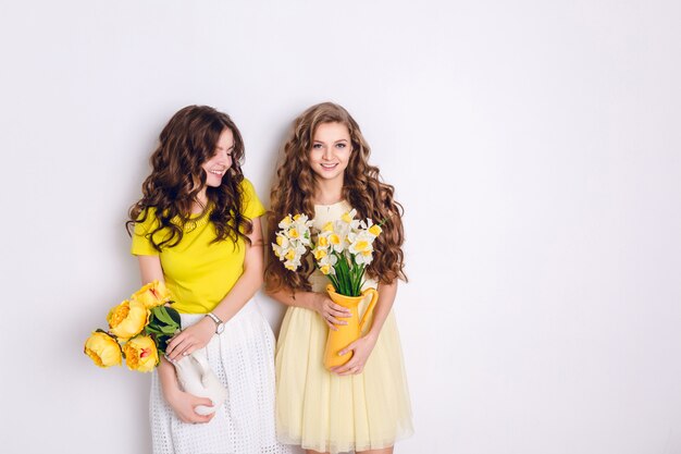 Foto de estudio de dos niñas sonrientes de pie. Una chica rubia y una chica morena sostienen jarrones con flores.