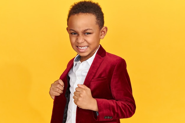 Foto de estudio de un elegante pequeño dandy de piel oscura que expresa verdaderas emociones, mostrando los dientes y haciendo muecas, habiendo indignado una mirada furiosa, apretando los puños, posando aislado en una chaqueta de terciopelo de moda