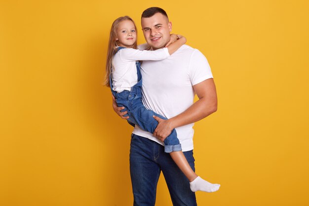 Foto de estudio de la encantadora niña niño con su padre, hombre guapo con niño en manos, vistiendo ropa casual