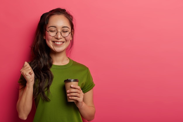 Foto gratuita foto de estudio de estudiante alegre levanta el puño, se regocija de triunfo y finaliza con éxito la tarea, tiene un descanso para tomar café después de largas horas de trabajo