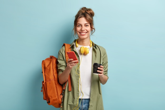 Foto de estudio de una estudiante alegre que toma un café después de las conferencias, escucha un audiolibro en auriculares, disfruta del registro del sitio web, usa el teléfono móvil para chatear en línea, tiene una mochila en la espalda.