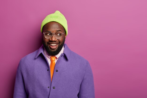 Foto de estudio de feliz hombre afroamericano negro mira con alegría a un lado, viste elegante chaqueta púrpura, sombrero y gafas ópticas, expresa emociones sinceras