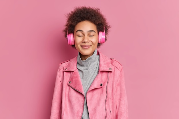 Foto de estudio de feliz joven afroamericana disfruta de una agradable melodía mantiene los ojos cerrados escucha música a través de auriculares