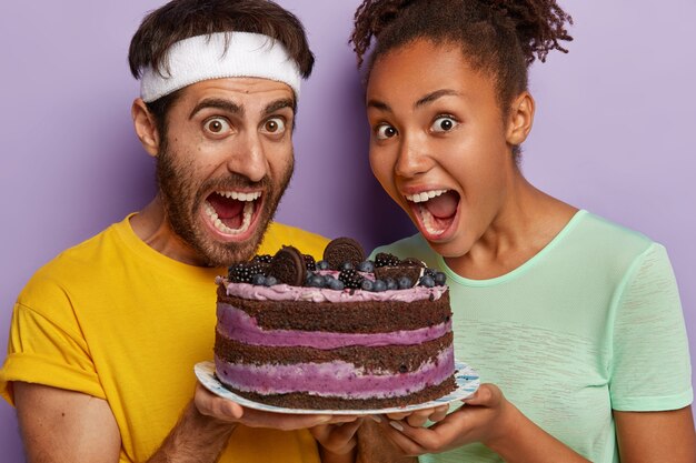 Foto de estudio de feliz pareja multiétnica llena de alegría mantenga delicioso pastel con arándanos