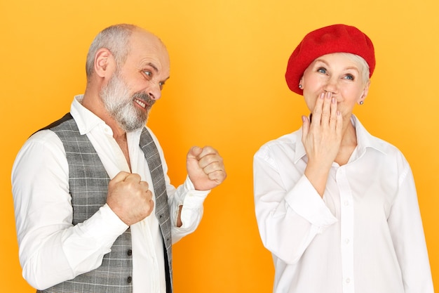 Foto de estudio de frívola despreocupada mujer de mediana edad en boina roja que cubre la boca jadeando, chico barbudo enojado apretando los puños, con mirada furiosa y loca, va a golpear a su esposa. Violencia doméstica