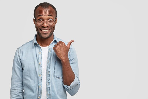 Foto de estudio de guapo alegre hombre de piel oscura con una gran sonrisa, indica con el pulgar a un lado