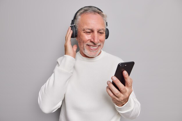 Foto de estudio de guapo senior usa gadgets modernos escucha música favorita a través de auriculares