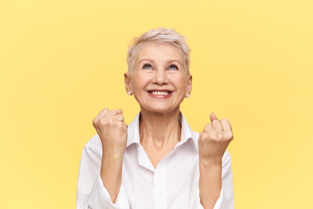 Foto de estudio de hermosa empresaria madura enérgica con peinado de duendecillo exclamando Sí, mirando hacia arriba con una sonrisa feliz, apretando los puños, celebrando un buen trato.