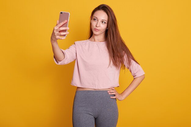 Foto de estudio de hermosa mujer de cabello castaño con cabello lacio en pantalones grises y camisa rosa tomando selfie