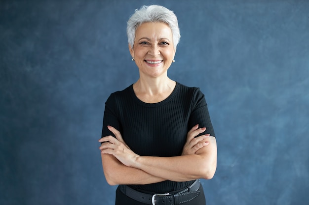 Foto de estudio de hermosa mujer caucásica jubilada feliz con peinado pixie cruzando los brazos sobre su pecho, con mirada de confianza, sonriendo ampliamente