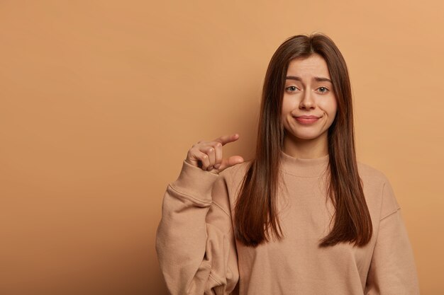 Foto de estudio de una hermosa mujer poco impresionada que hace un gesto de pequeño tamaño, demuestra un objeto pequeño, usa una sudadera suelta