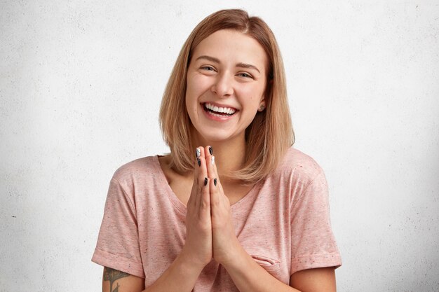 Foto de estudio de una hermosa mujer sonriente que mantiene la palma junta, tiene una expresión alegre, le pide al esposo que compre su nuevo atuendo, estando de buen humor