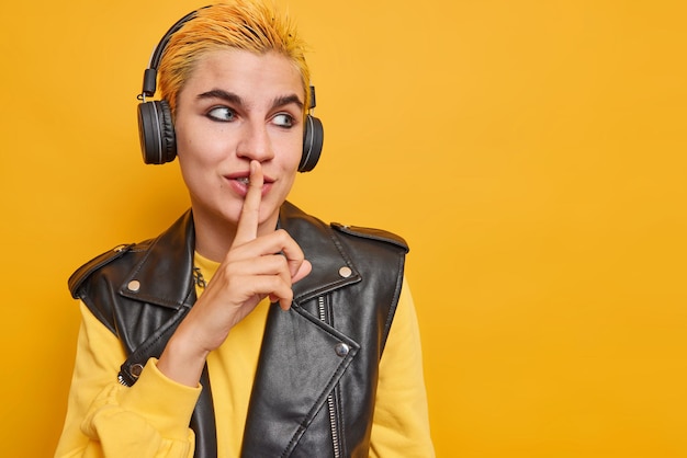 Foto de estudio de hister pensativa chica hace gesto de silencio mantiene el dedo índice en los labios dice información secreta concentrada en el espacio en blanco escucha música en auriculares usa ropa elegante