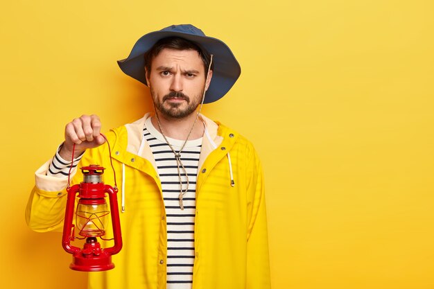 Foto de estudio de un hombre de descontento serio que tiene poca barba y bigote, listo para descubrir un nuevo lugar en la oscuridad, sostiene una lámpara de queroseno, vestida con un impermeable y un oído.