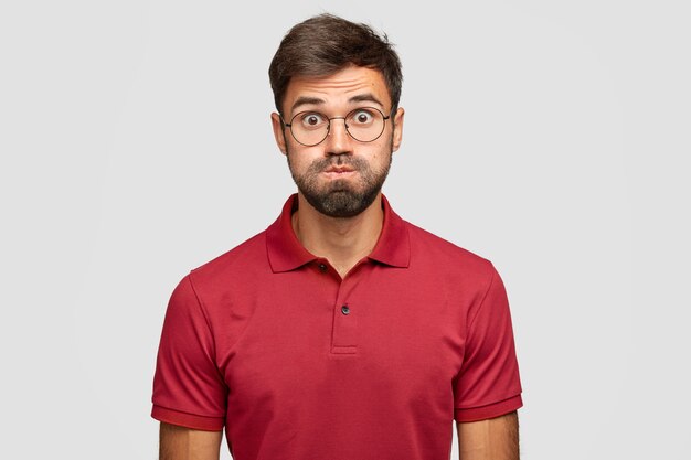 Foto de estudio de hombre emotivo ha disgustado la expresión, sopla las mejillas, tiene los ojos abiertos