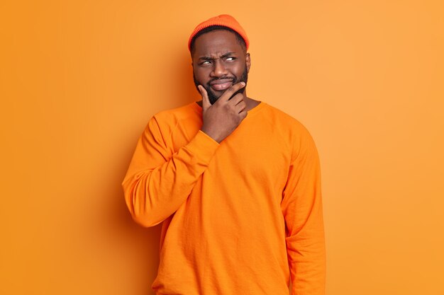 Foto de estudio de hombre guapo con barba sostiene la barbilla mira pensativamente a un lado piensa profundamente sobre algo lleva sombrero y poses de suéter contra la pared de color naranja vivo brillante