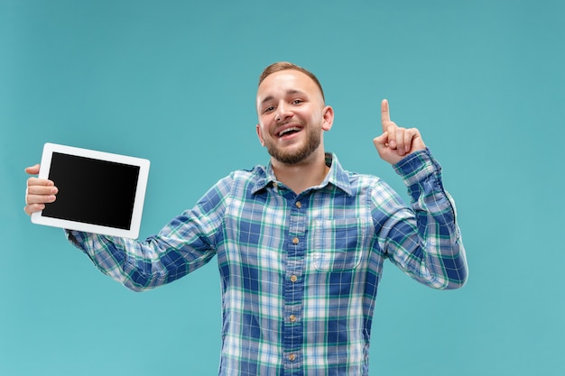 Foto de estudio del hombre positivo aislado en la pared azul de pie en ropa casual con tableta y mostrándolo en blanco con una sonrisa feliz