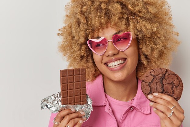 Foto de estudio de una joven alegre que tiene un gusto por lo dulce, sostiene una barra de chocolate y una galleta elige entre dos postres, las sonrisas en general usan gafas de sol rosas y una chaqueta aislada sobre fondo blanco