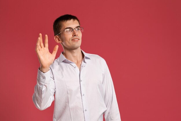 Foto de estudio de un joven elegante con una camisa blanca clásica, relojes negros y anteojos saludando a alguien mientras posa sobre un fondo rosa. corte de pelo con estilo. concepto de emociones sinceras. copia