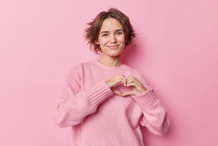 Foto gratuito una foto de estudio de una joven europea feliz hace un gesto de corazón sobre el pecho expresa sonrisas de amor y usa un saltador casual aislado sobre un fondo rosado. chica alegre comparte sentimientos sinceros