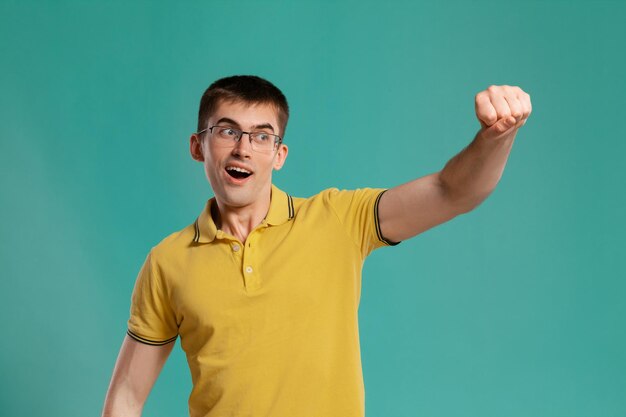 Foto de estudio de un joven interesante con una camiseta informal amarilla, anteojos y relojes negros que se ven muy felices mientras posan sobre un fondo azul. Corte de pelo con estilo. Concepto de emociones sinceras. Copiar sp