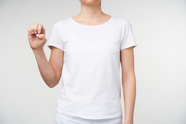 Foto de estudio de una joven de piel clara aprendiendo el alfabeto sordo y formando la letra e con la mano levantada mientras está de pie sobre un fondo blanco en ropa casual