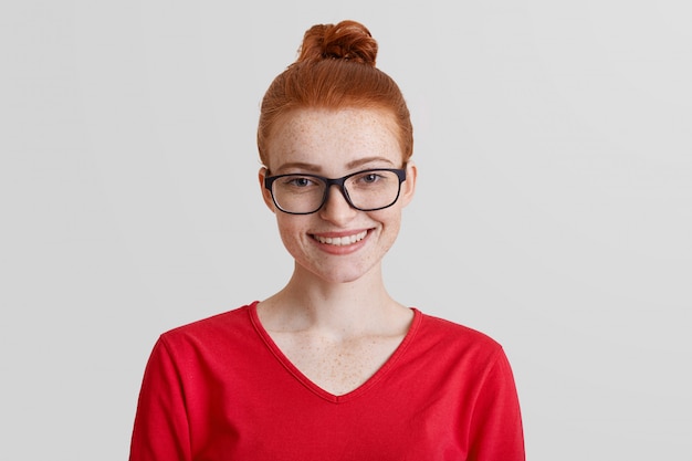 Foto de estudio de una joven sonriente pelirroja que usa gafas cuadradas y un suéter rojo, que está de buen humor después de la promoción en el trabajo, recibe un premio por su trabajo diligente y logra un gran éxito