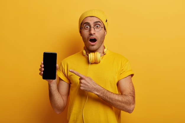 Foto de estudio de un joven sorprendido apunta a la pantalla del teléfono inteligente, muestra una pantalla en negro, demuestra un producto moderno