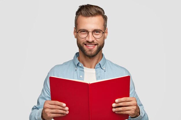 Foto de estudio de lector hombre alegre con expresión satisfecha, sostiene el libro rojo