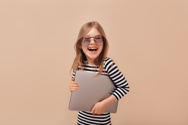 Foto de estudio de una linda chica sonriente feliz con gafas de moda y sosteniendo un portátil sobre fondo beige