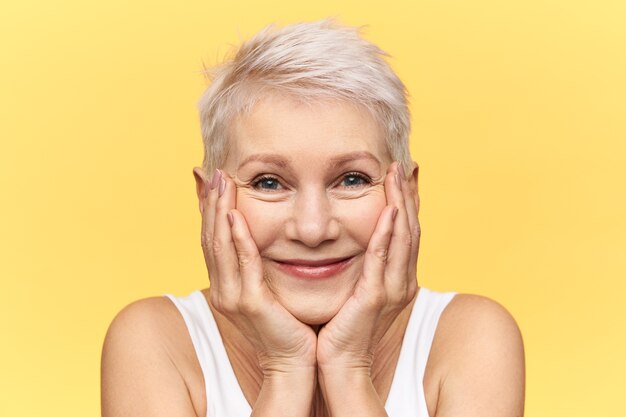 Foto de estudio de linda divertida mujer de mediana edad con el pelo corto teñido sonriendo, colocando las mejillas en las manos, mirando a la cámara con expresión facial curiosa feliz, viendo algo muy interesante