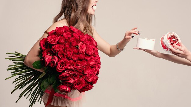 Foto de estudio de una linda mujer morena sosteniendo un ramo de hermosas rosas rojas. Está comiendo dulces y tomando un caramelo más de la caja en manos femeninas anónimas. Aislada en blanco.