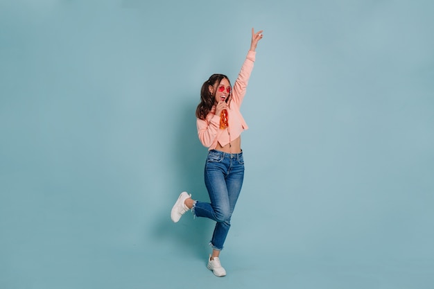 Foto de estudio de longitud completa de una joven atractiva vestida con suéter rosa y gafas redondas rosas bailando y bebiendo jugo sobre una pared aislada