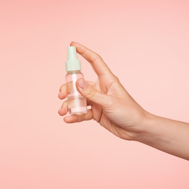 Foto de estudio de la mano de piel clara de la mujer joven manteniendo el dedo índice en el botón de la botella mientras va a rociar contenido, aislado en rosa