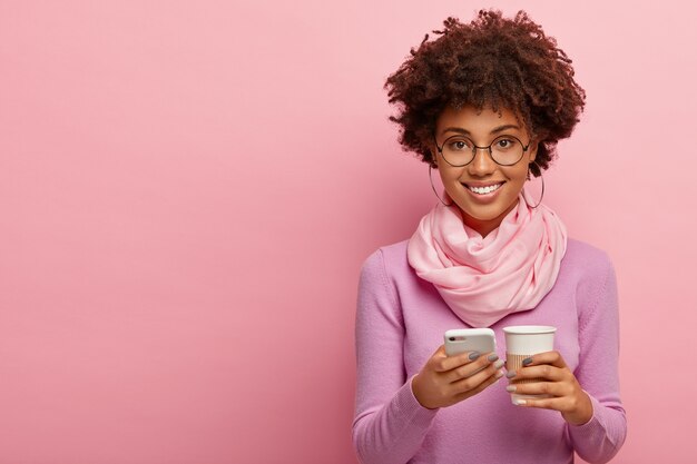 Foto de estudio de una mujer afroamericana encantada que navega por Internet en un teléfono inteligente, revisa las noticias, disfruta bebiendo café aromático en un vaso de papel, usa ropa de moda y gafas redondas