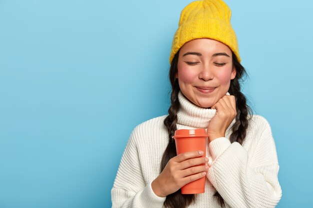 Foto de estudio de una mujer asiática de aspecto agradable que tiene dos trenzas, viste un sombrero amarillo y un suéter blanco de gran tamaño, sostiene café para llevar, posa sobre un fondo azul, copia espacio para su publicidad