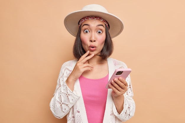 Foto de estudio de una mujer asiática sorprendida que viste ropa elegante de fedora tiene una cara muy sorprendida recibe un mensaje inesperado o un comentario debajo de su publicación en las redes sociales sostiene que el teléfono móvil usa internet