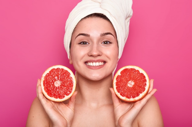 Foto de estudio de mujer atractiva con pomelo en sus manos y con una toalla blanca sobre su cabeza, mujer después de tomar una ducha o baño, estar de buen humor, posando con una sonrisa de dientes. Concepto de cuidado de la piel.