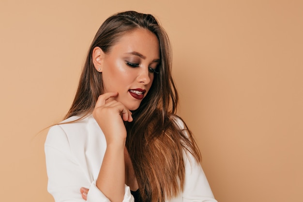 Foto de estudio de mujer bonita europea con maquillaje brillante mirando hacia abajo con una sonrisa sobre fondo