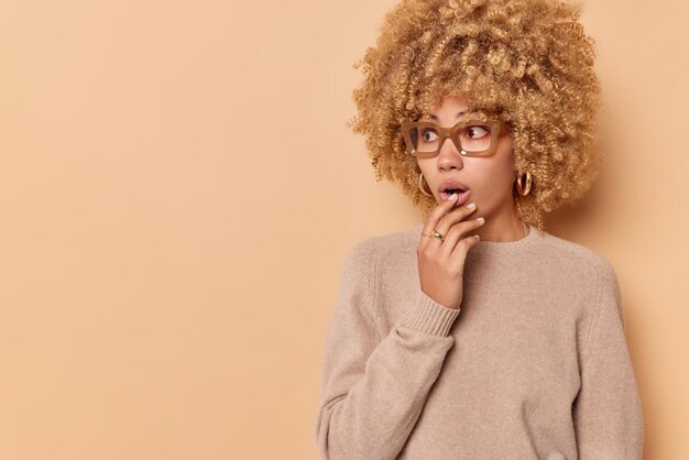 Una foto de estudio de una mujer de cabello rizado impresionada se ve asombrada y mantiene la boca abierta reacciona ante noticias impactantes usa anteojos y un puente casual de manga larga aislado sobre el espacio de copia de fondo marrón