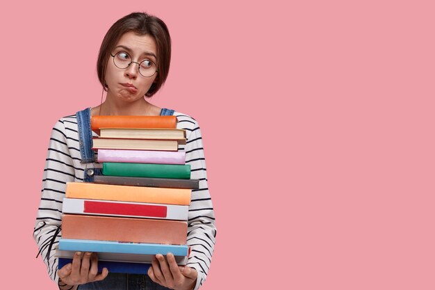 Foto de estudio de mujer caucásica insatisfecha muerde los labios, usa gafas y un jersey de rayas, no quiere estudiar