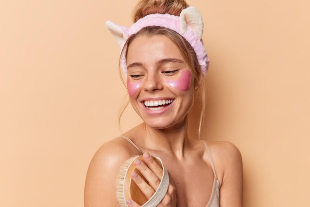 Foto de estudio de una mujer feliz que se lava el cuerpo con un cepillo de baño, sonríe alegremente posa sin camisa, usa diadema, aplica parches de hidrogel debajo de los ojos aislados sobre fondo beige. Concepto de cuidado de la piel facial