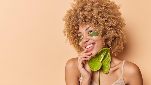 Foto gratuita foto de estudio de una mujer guapa con cabello rizado y tupido aplica parches cosméticos en los ojos sostiene calla sonríe alegremente se encuentra con los hombros desnudos aislados sobre el espacio de copia de fondo marrón para su texto