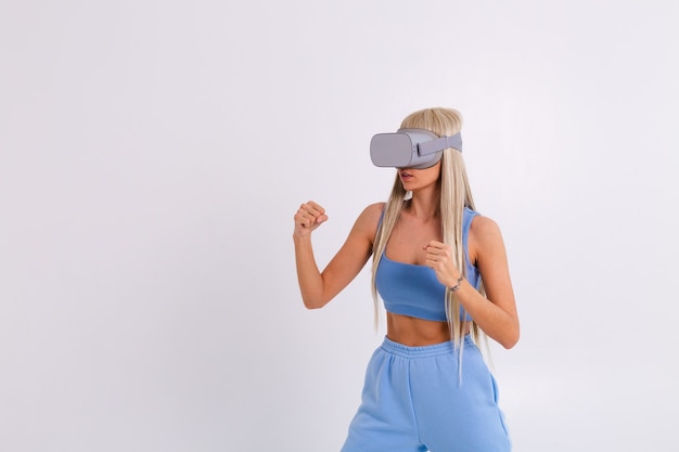 Foto de estudio de una mujer joven y atractiva en un traje de moda azul cálido con gafas de realidad virtual sobre un fondo blanco juega una pelea de boxeo