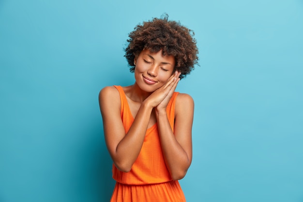 Foto de estudio de una mujer joven y bonita que se inclina sobre las palmas presionadas, cierra los ojos y tiene una sonrisa agradable, sueña con algo vestido con un vestido de verano naranja aislado en la pared azul del estudio