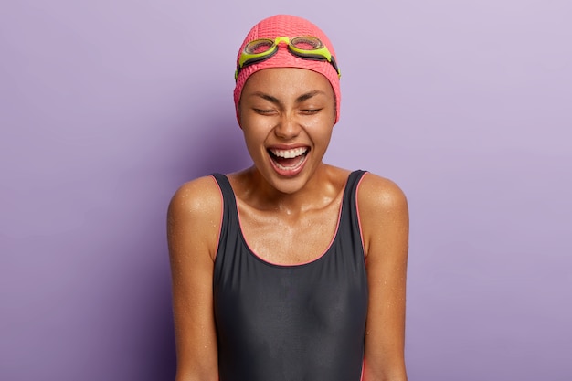 Foto de estudio de mujer de piel oscura llena de alegría cierra los ojos, vestida con traje de baño, gafas en la cabeza, pasa el tiempo libre en el centro deportivo, tiene el cuerpo mojado, feliz de terminar la carrera. Buceo y ejercicio