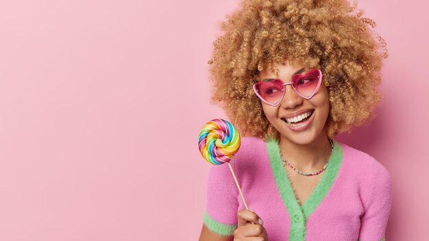 Una foto de estudio de una mujer positiva con sonrisas de cabello rizado usa gafas de sol en forma de corazón y una camiseta sostiene dulces coloridos aislados sobre un espacio en blanco de fondo rosa para su contenido publicitario