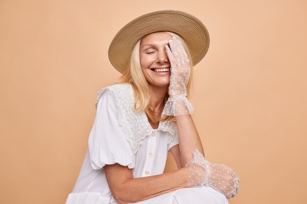 Foto de estudio de una mujer rubia alegre y despreocupada que hace que la palma de la cara se ría felizmente de una buena broma viste un vestido blanco de moda y guantes de encaje se sienta contra la pared beige