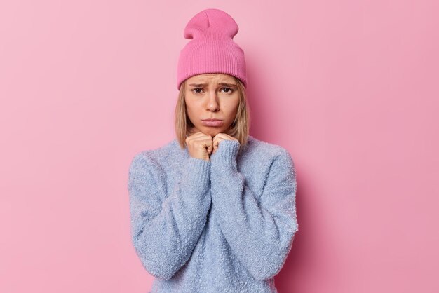 Una foto de estudio de una mujer triste disgustada que mantiene las manos bajo la barbilla se siente infeliz vestida con un suéter casual y un sombrero aislado sobre un fondo rosa Una modelo femenina frustrada y malhumorada expresa emociones negativas