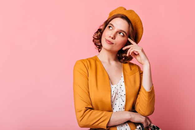 Foto de estudio de pensativa chica francesa mirando hacia arriba. Encantadora mujer joven en boina pensando en algo sobre fondo rosa.